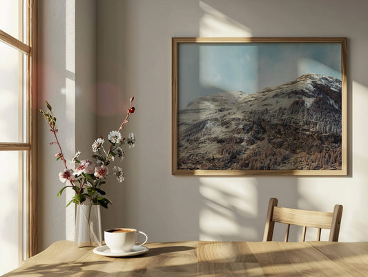 Analog fine art mountain landscape print of alpine peaks between seasons, shot on 35mm film in the Swiss Alps, displayed in a wooden frame in a sunlit minimalist dining interior