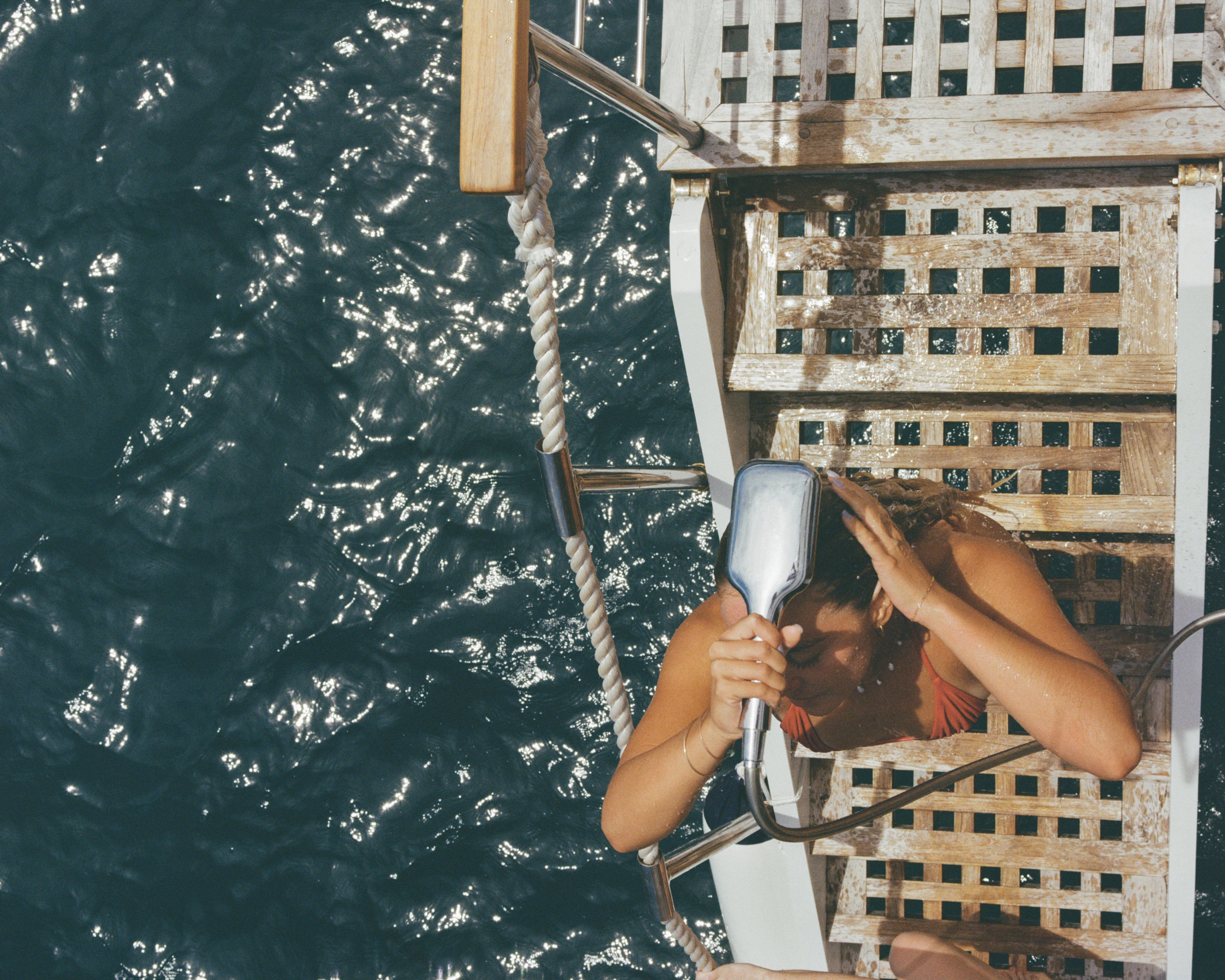 Analog photograph of woman rinsing her hair on a boat ladder above deep blue water, sunlight reflecting on the surface, shot on medium format film