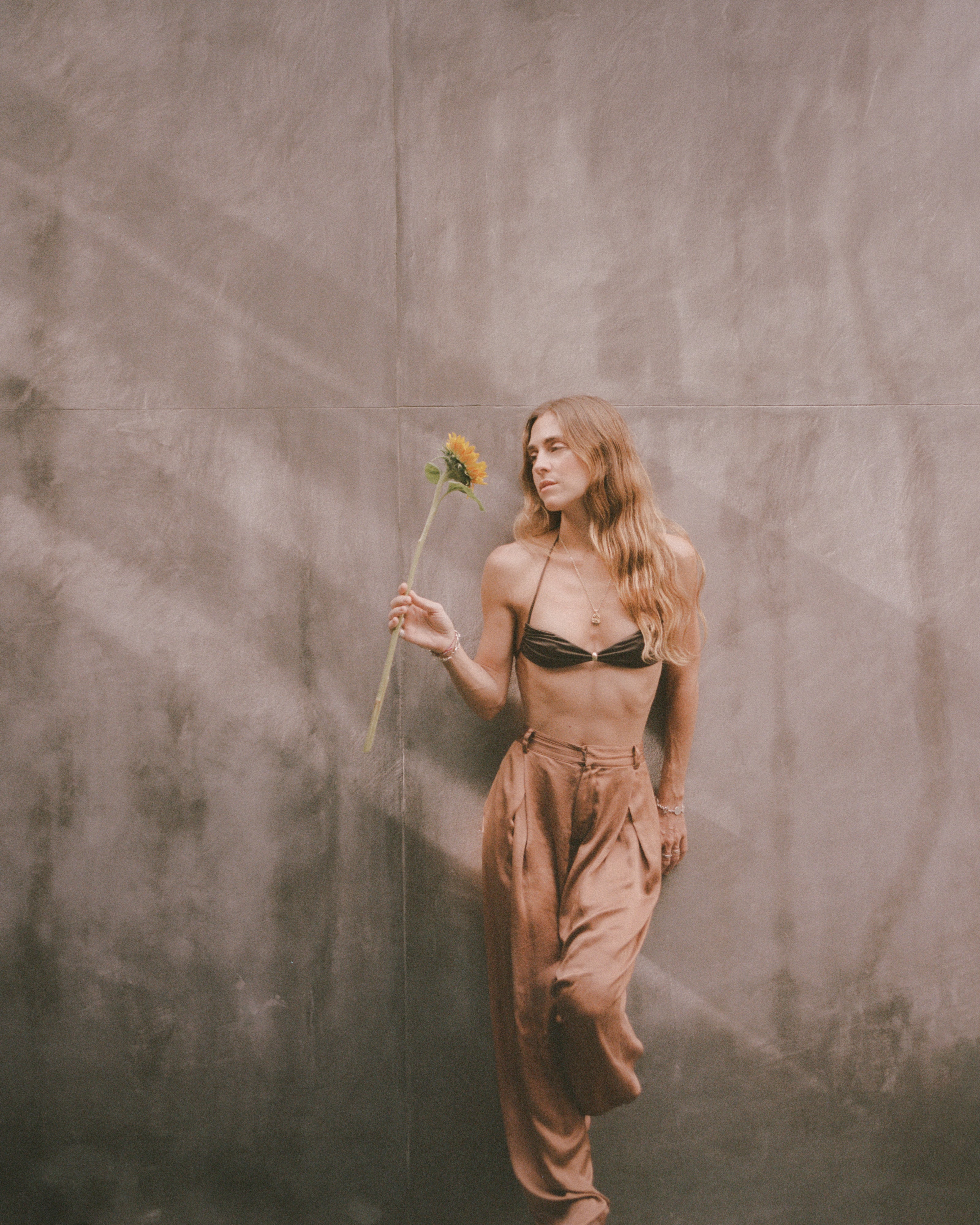 Analog fashion portrait of model holding a sunflower against a textured wall, soft natural light and muted tones, shot on medium format film