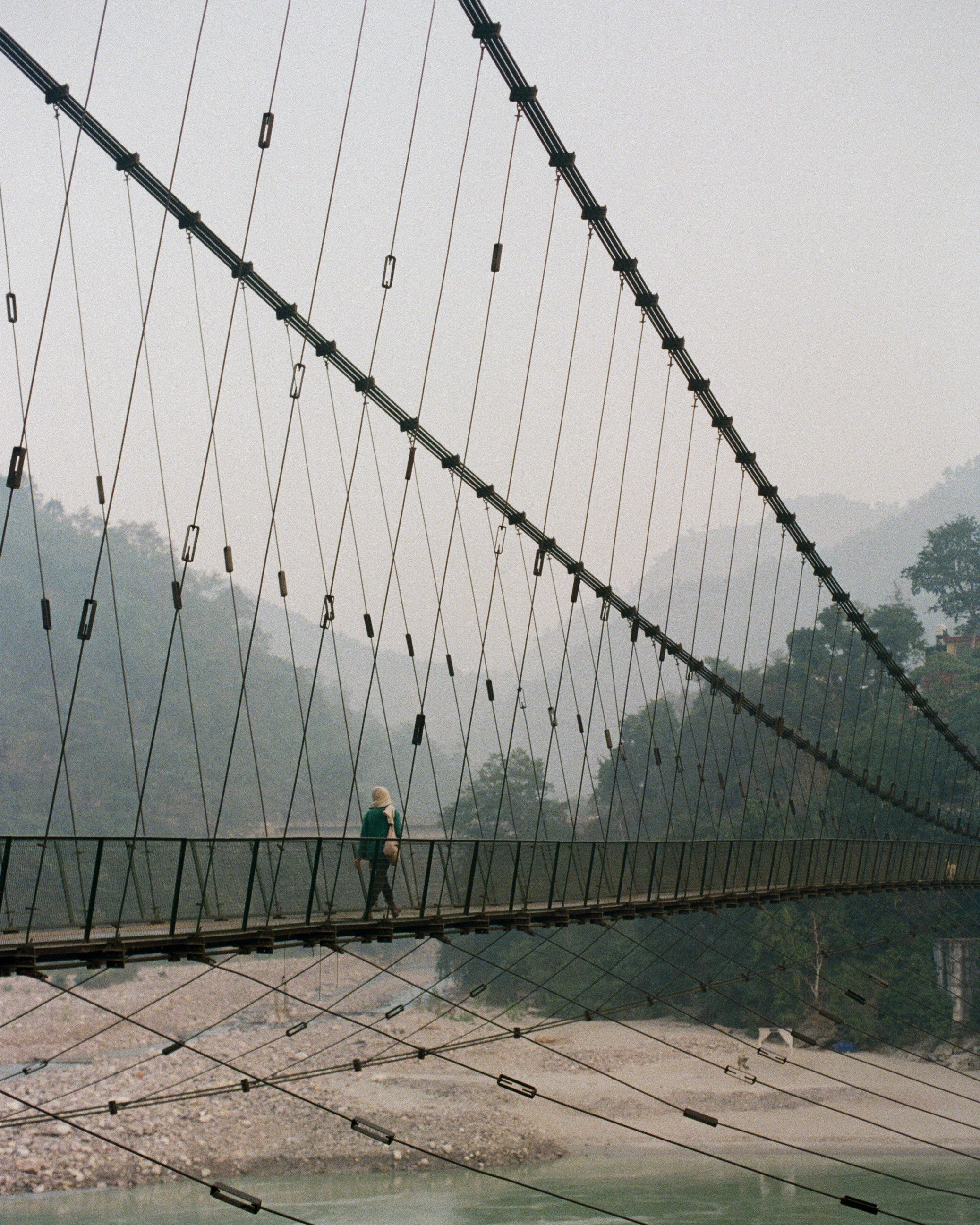 Analog photograph of solitary figure walking across suspension bridge above river with misty mountains in the background, shot on medium format film