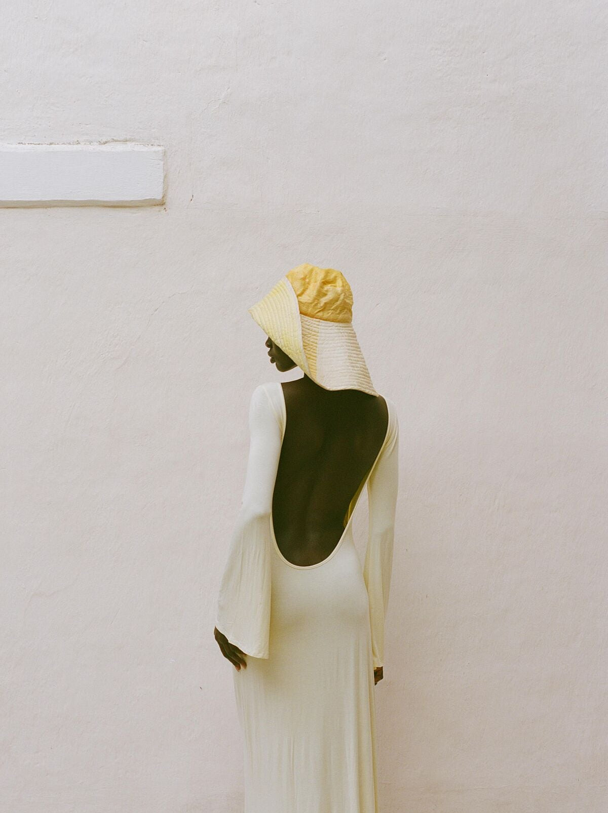 Analog fashion portrait of woman in open back cream dress and straw hat shot on 35mm film against minimalist white wall in warm natural light.