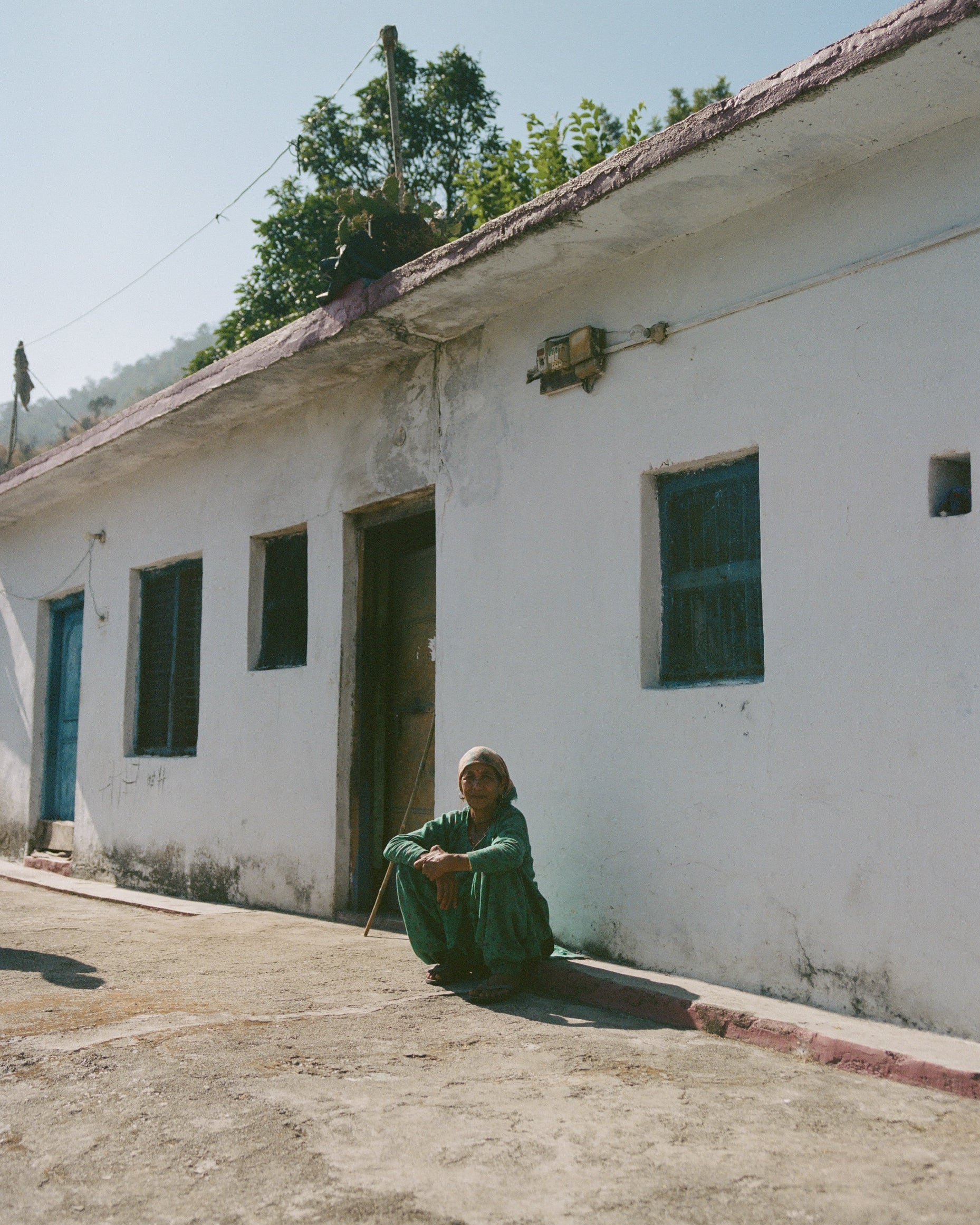 Analog documentary photograph of local woman seated outside a white house in the Indian mountains, captured in natural sunlight on medium format film