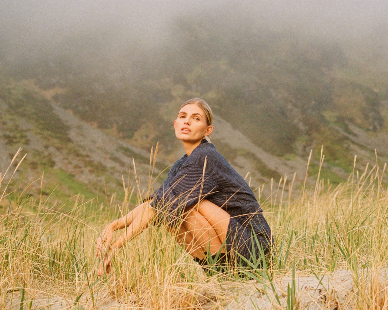 Analog fashion portrait of Louise seated in tall grass in a misty landscape in Norway, shot on film in soft natural light