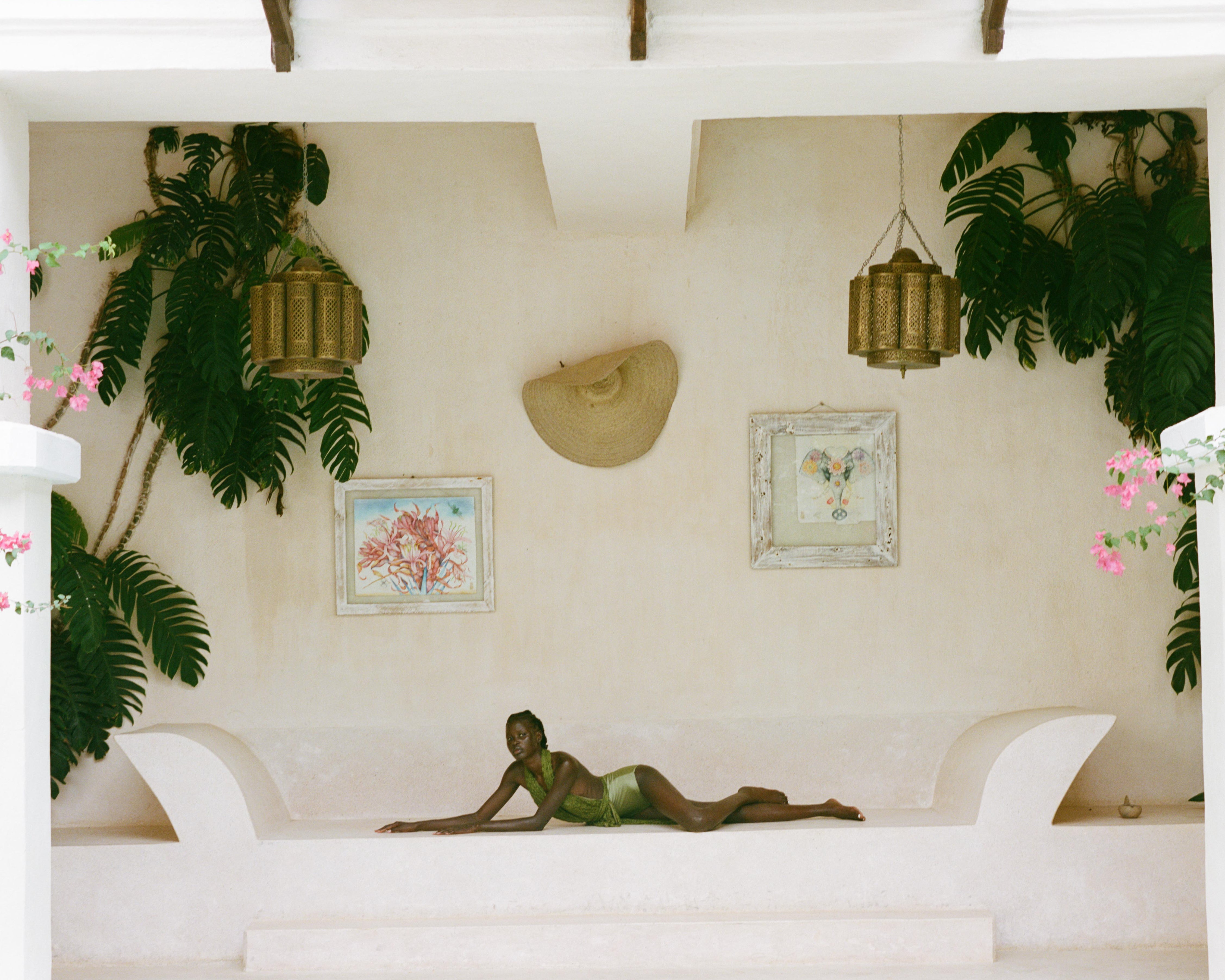 Analog fashion image of model reclining on a built-in bench within a minimal interior with plants, artwork, and hanging lanterns, shot on medium format film