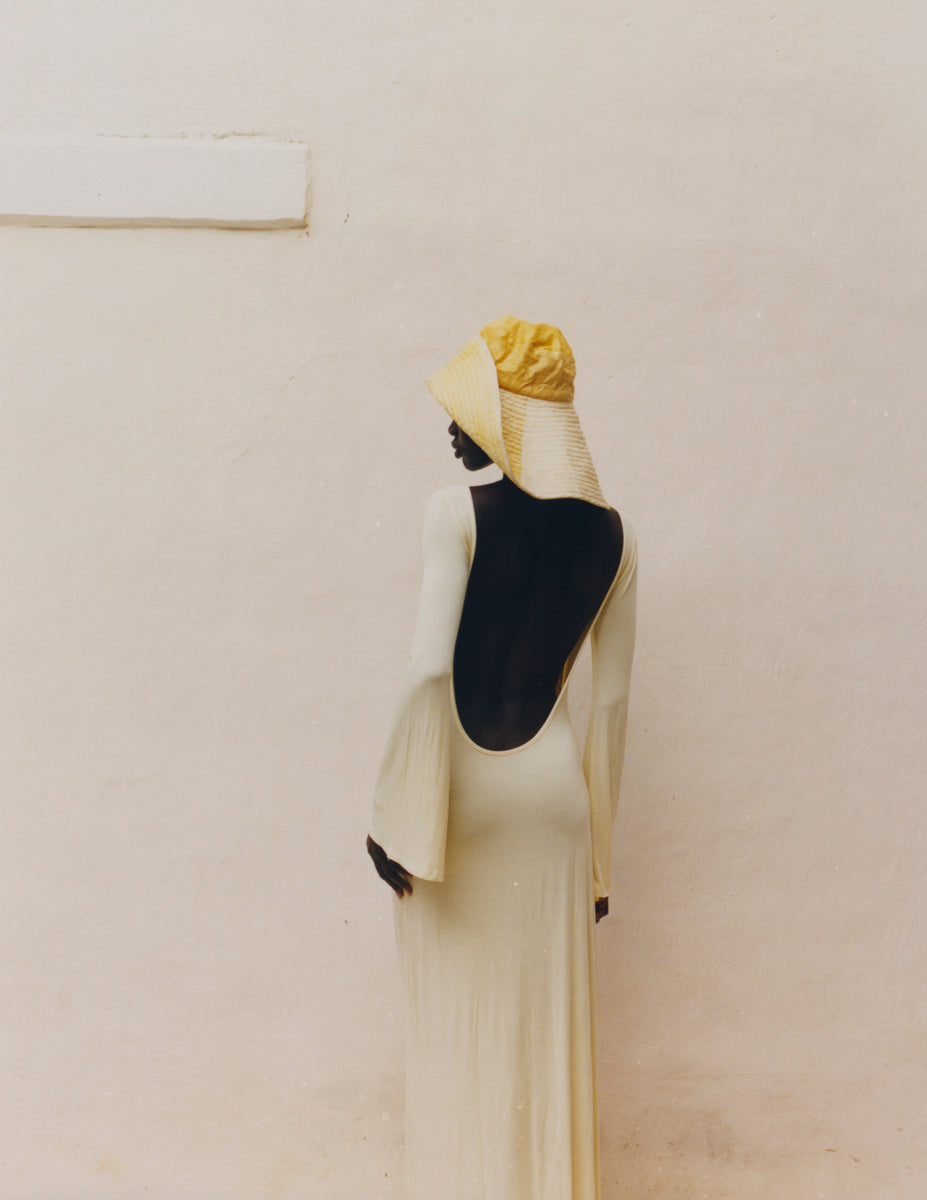 Analog fashion portrait of woman in open back cream dress and straw hat shot on 35mm film against minimalist white wall in warm natural light