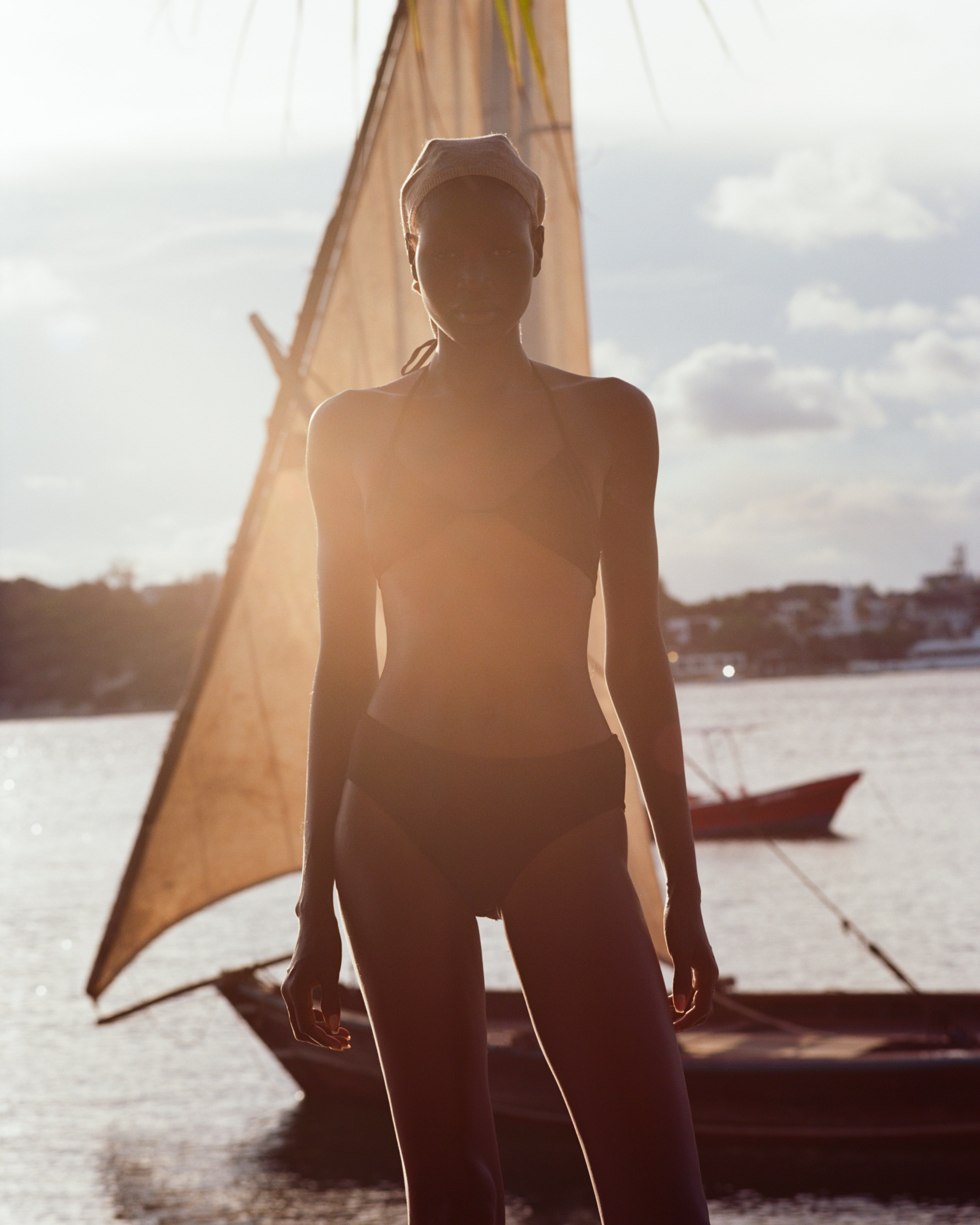 Analog fashion portrait of model wearing OAD swimwear, backlit by warm sunset light with dhow boats in the background, shot on medium format film