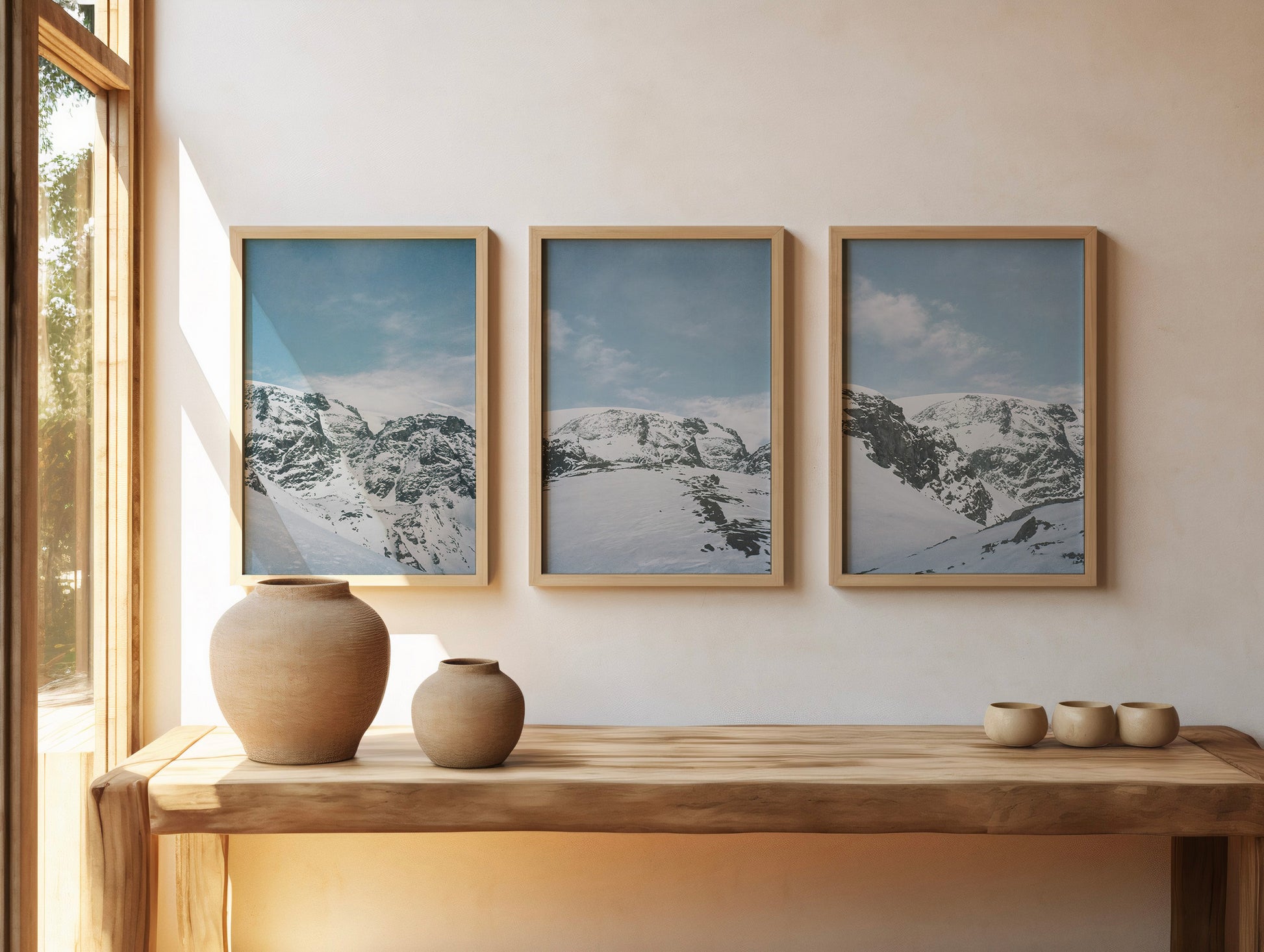 Minimalist interior with three framed snow-covered mountain landscape prints in soft blue tones, styled above a wooden console with ceramic vases and natural light, fine art analog photography print shot on 35mm film