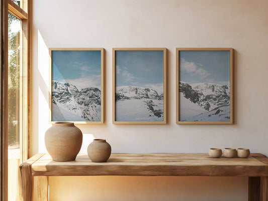 Minimalist interior with three framed snow-covered mountain landscape prints in soft blue tones, styled above a wooden console with ceramic vases and natural light, fine art analog photography print shot on 35mm film