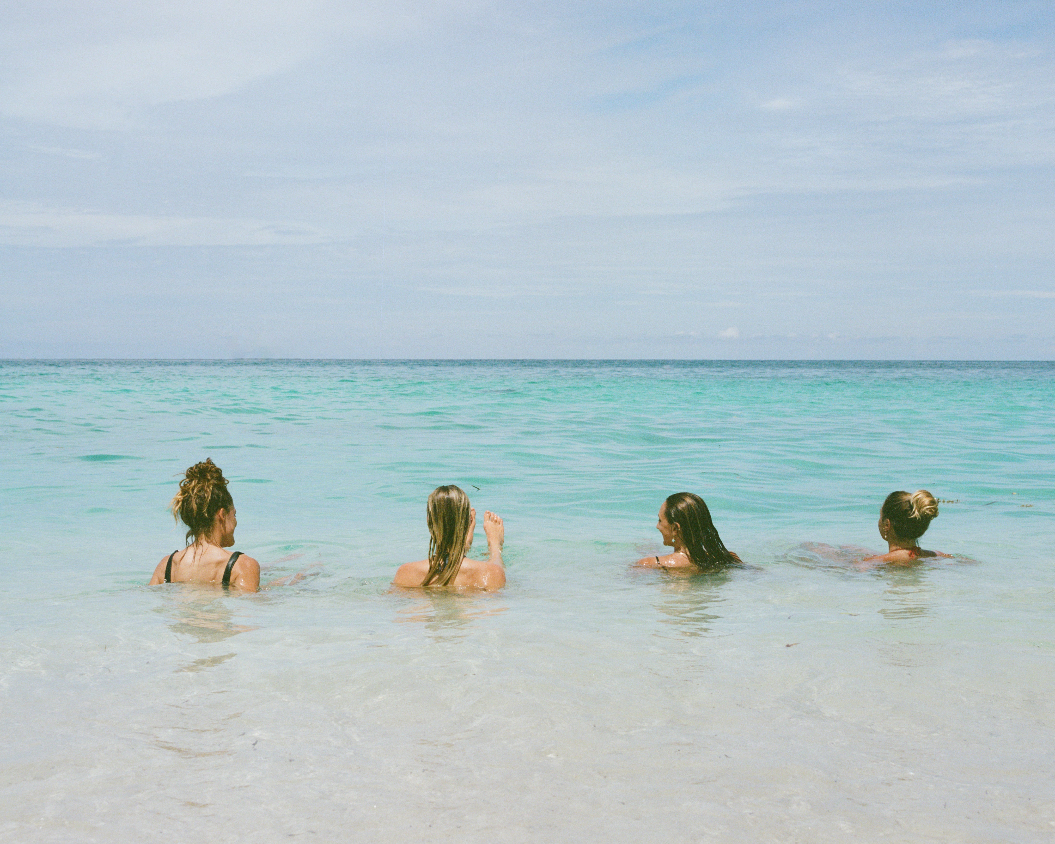 Analog photograph of four women swimming in shallow turquoise ocean, facing the horizon under soft sky, shot on medium format film