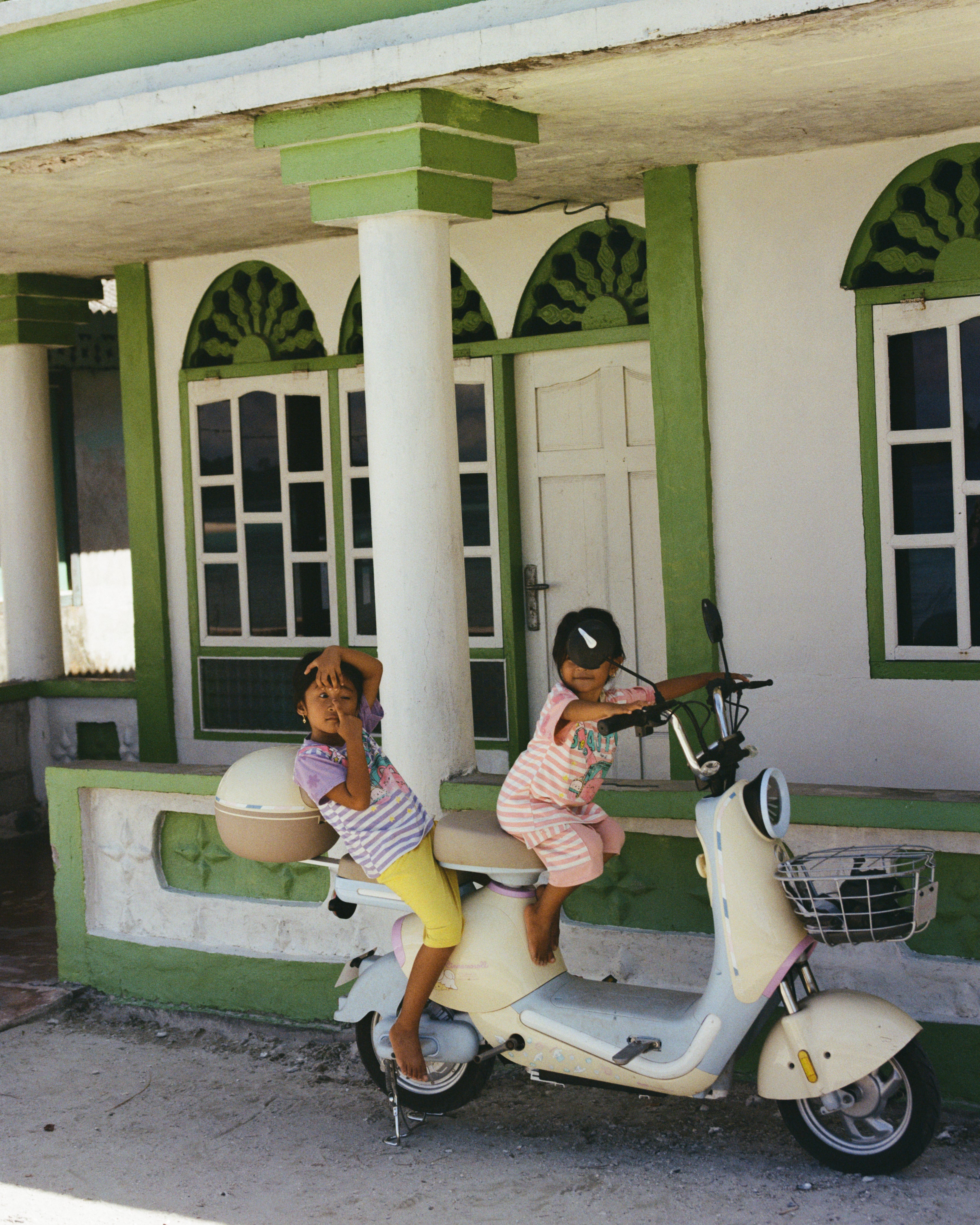 Two children riding a scooter in front of a house with green and white exterior. shot in mentawais on remote island in indonesia on 35mm. 
