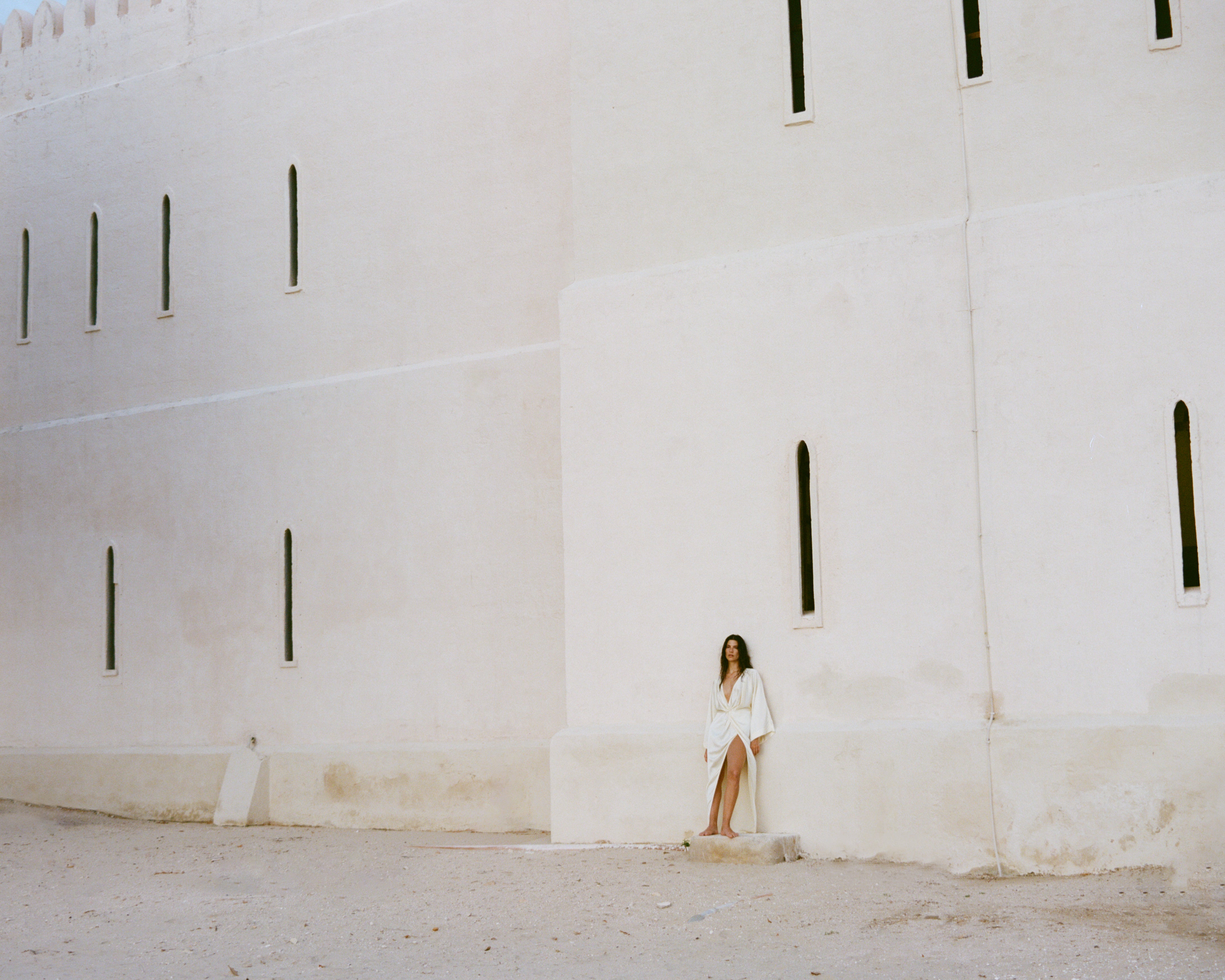 Woman in a white dress sitting against a white wall with vertical black slits. Fort Shala Lamu Kenya
